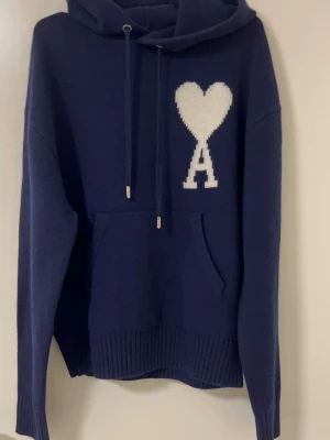 Mörkblå hoodie från Ami Paris - Mörkblå hoodie från Ami Paris med stor vit hjärtlogga och bokstaven A på bröstet. Hoodien har huva med dragsko, magficka och ribbade muddar vid ärmslut och nederkant. Tillverkad i mjukt stickat material som känns lyxigt och bekvämt. I ny skick, knappt använd 