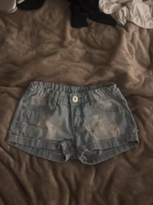 Ljusblå denimshorts med slitningar - Säljer ett par ljusblå denimshorts med slitna detaljer och upprullade benslut. Klassisk femficksmodell med knapp och dragkedja framtill. Perfekta för varma dagar och en avslappnad stil.