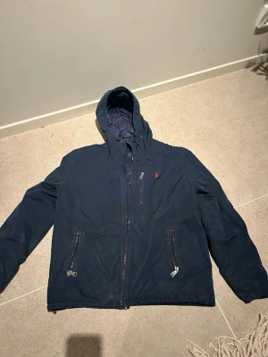 Marinblå vindjacka från Polo Ralph Lauren - Snygg marinblå vindjacka från Polo Ralph Lauren med huva och dragkedja framtill. Jackan har två framfickor med dragkedja och den klassiska röda loggan på bröstet. Tillverkad i ett lätt och slitstarkt material, perfekt för blåsiga dagar.