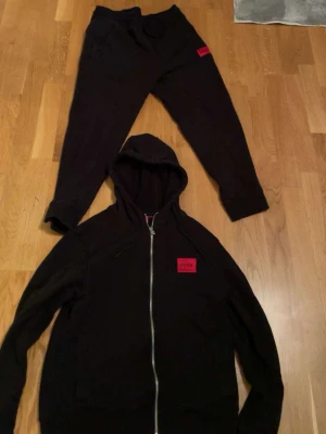 Svart HUGO loungewear set - Säljer ett svart loungewear set från HUGO med matchande hoodie och byxor. Hoodien har dragkedja, huva och röd HUGO-logga på bröstet. Byxorna har resår i midjan och röd logga på benet. Perfekt för chill dagar hemma.