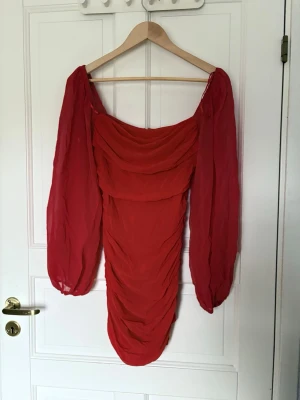 Röd kort klänning - Trendig röd kortklänning med långa, transparenta puffärmar. Klänningen har draperad effekt framtill och baktill samt en smickrande, figurnära passform. Perfekt för dig som vill sticka ut med en färgstark och modern look.