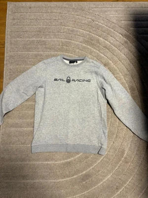 Grå sweatshirt från Sail Racing - Snygg grå sweatshirt från Sail Racing med svart logotyptryck på bröstet. Tröjan har rund halsringning, långa ärmar och ribbade muddar vid ärmslut och nederkant. Perfekt för en avslappnad och sportig stil.