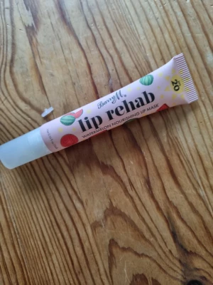 Barry M Lip Rehab Watermelon läppmask - Säljer en Barry M Lip Rehab Watermelon Nourishing Lip Mask i tub. Läppmasken har en ljusrosa förpackning med färgglada vattenmelonmotiv och gul text. Innehåller vårdande ingredienser som kakaosmör och sheasmör samt SPF 20. Perfekt för att återfukta och skydda läpparna.