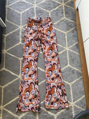 Färgglada retro-m/g mönstrade leggings - Säljer ett par riktigt färgglada leggings med retroinspirerat blommönster i orange, rosa, brunt och blått. De har utsvängda ben och elastisk midja för skön passform. Perfekta för dig som vill sticka ut med en unik och lekfull stil!