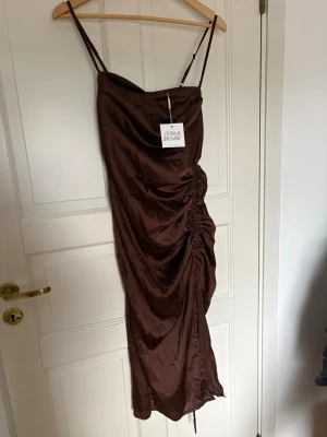 Brun omlottklänning Public Desire - Snygg brun långklänning från Public Desire med smala axelband och omlottdesign. Klänningen har en draperad effekt på sidan och är tillverkad i ett glansigt material.
