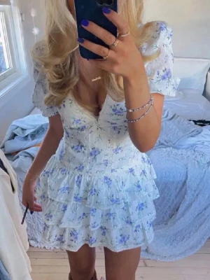 Blommig kortklänning med volanger - Supersöt kortklänning med puffärmar och v-ringning. Klänningen är vit med blått och lila blommönster. Inga skador