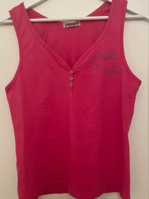 Rosa linne från Onda D'oro - Snyggt rosa linne från Onda D'oro med V-ringning och tre små knappar framtill. Dekorerad med texten 'Onda D'oro' i glittriga stenar på bröstet. Tillverkat i mjukt bomullsmaterial och har breda axelband för en bekväm passform. Super fin till sommaren eller när man vill vara lite extra färgglad! Har en underbar vintage, y2k utseende 🩷🩷🩷