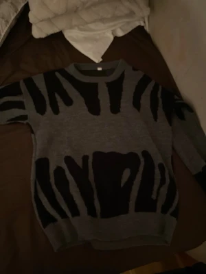 Två stickade tröjor med mönster - Säljer två stickade tröjor med coola mönster. Ena är grå med svart zebrainspirerat mönster och rund hals, den andra är ljusgrå med abstrakt mönster i mörkgrått. Båda har långa ärmar och ribbade muddar. Perfekta för dig som gillar unika stickade plagg.