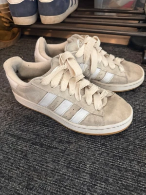 Beige Adidas sneakers med mocka - Snygga beige sneakers från Adidas i mocka med klassiska vita ränder på sidan och vit sula. De är i storlek 40 och dem har inte hål någonstans eller sönder.   Skorna har breda vita skosnören och låg profil som ger en clean och stilren look man får även med helt nya vita skosnören som var med i paketet när jag köpte dem. Perfekta för dig som gillar retrovibbar och enkel design.