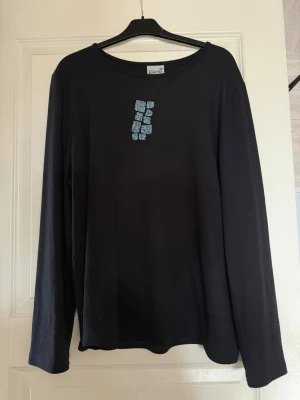Svart långärmad topp med tryck - Svart långärmad topp från Klimt med ett unikt blått tryck framtill på bröstet. Toppen har en avslappnad passform och rund halsringning. Perfekt att styla med både jeans och kjol för en cool look.