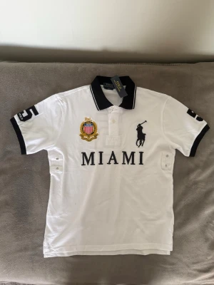 Chief Keef Polo Ralph Lauren Miami piketröja - Vit pikétröja från Polo Ralph Lauren med svart krage och detaljer. Stor broderad logga på bröstet, MIAMI-tryck och emblem. Svarta detaljer på ärmarna och klassisk pikéknäppning. Perfekt för en sportig och stilren look. PRIS KAN DISKUTERAS