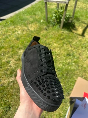 Svarta spikes-sneakers från Louboutin - Snygga svarta sneakers från Christian Louboutin med mattsvarta nitar på tån, svart mocka och klassisk röd sula med logga. Skorna har svarta skosnören och en låg siluett som ger en edgy och exklusiv look.