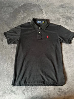 Svart pikétröja från Ralph Lauren - Säljer en klassisk och stilren pikétröja från Polo Ralph Lauren i svart med den ikoniska röda loggan på bröstet. Modellen är Custom Slim Fit, vilket ger en modern och mycket snygg passform.  Märke: Polo Ralph Lauren Storlek: M (Custom Slim Fit) Färg: Svart med röd broderad logga Skick: I absolut nyskick! Endast använd 3 gånger och mycket väl omhändertagen. Inga blekningar, hål eller fläckar.  📦 Frakt: Jag skickar samma dag eller senast dagen efter betalning med spårbar frakt.