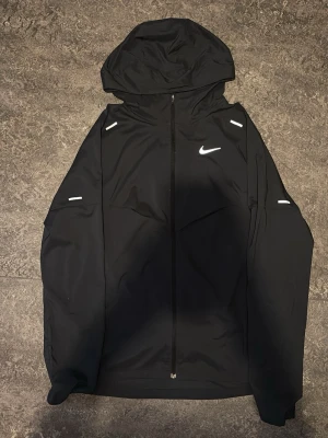 Svart Nike vindjacka med huva - Svart vindjacka från Nike med hel dragkedja, huva och reflekterande detaljer på axlar och ärmar. Jackan har en minimalistisk design med Nike-logga på bröstet och är tillverkad i lätt, vindtätt material. Perfekt för blåsiga dagar och sportiga outfits.