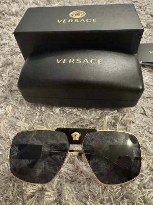  solglasögon Versace - Snygga svarta solglasögon från Versace med fyrkantig form och gulddetaljer. Klassisk Medusa-logga i guld på båda sidor och över näsryggen. Glasögonen har en exklusiv känsla med svarta och guldiga detaljer, och levereras med originaletui och ask.