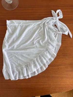 Vit strandkjol med volang och knytband - Säljer en vit strandkjol i lätt och luftig textil från Shein. Kjolen har volangkant längs nederdelen och knyts i sidan för en snygg och justerbar passform. Perfekt att slänga på sig över bikinin på stranden. Materialet känns mjukt och svalt. Passar strl S-M.