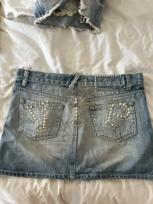 Blå jeanskjol med pärldetaljer - Säljer en kort jeanskjol i ljusblå denim med pärldetaljer på fickor och linning. Kjolen har klassisk femficksmodell, bälteshällor och stängs med knapp och dragkedja. Perfekt för dig som vill ha en unik och trendig look. Midjemått rakt över: 37cm. Längd: 31cm. Den är lite smutsig (sista bilden) men går säkert bort i tvätten. 