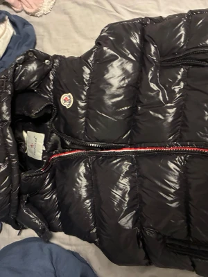 Svart dunväst från Moncler - Snygg svart dunväst från Moncler med glansig finish och klassisk Moncler-logga på bröstet. Västen har dragkedja framtill med röd, vit och blå detalj samt praktiska fickor med dragkedja. Perfekt för lager-på-lager och streetstyle.