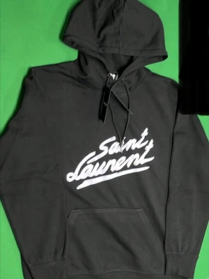 Svart Saint Laurent hoodie - Snygg svart hoodie från Saint Laurent med stor vit logga i graffiti-stil på bröstet. Hoodien har huva med dragsko och en klassisk magficka. Tillverkad i mjukt material som känns skönt mot huden. Perfekt för dig som vill ha en streetwear-look.