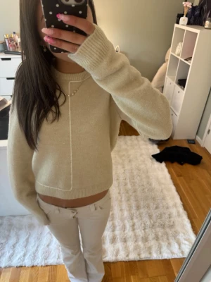Beige stickad tröja med ribbade muddar - Mysig beige stickad tröja med rund hals och ribbade muddar vid ärmslut och nederkant. Tröjan har en enkel, rak passform och en diskret sömdetalj framtill. Perfekt för lager på lager och passar till det mesta.