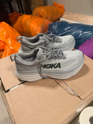 Hoka Bondi 8 grå sneakers - Snygga Hoka Bondi 8 sneakers i ljusgrått med svart logga på sidan. Ovandelen är i mesh för bra ventilation och sulan är extra tjock för maxad komfort. Perfekta för dig som gillar streetwear och vill ha riktigt sköna skor till vardags.