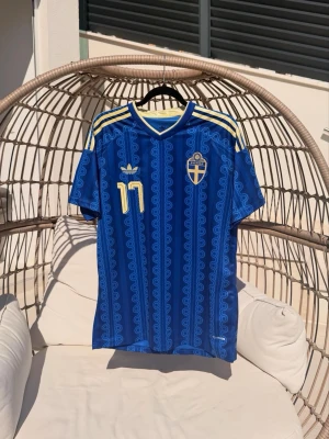 Blå Sverige matchtröja Adidas - Säljer en blå Sverige matchtröja från Adidas med gula detaljer och nummer 11 på bröstet. Tröjan har korta ärmar, V-ringning och det svenska landslagets emblem på vänster sida. Tillverkad i lätt och ventilerande material, perfekt för fotboll eller träning.