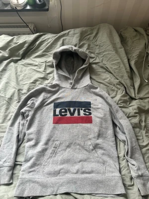 Grå hoodie från Levi's med logga - Säljer en grå hoodie från Levi's med stor färgglad logga på bröstet i blått, vitt och rött. Hoodien har huva och en klassisk magficka. Tillverkad i mjukt sweatshirtmaterial som är perfekt för chill dagar.
