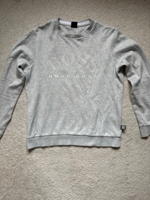 Grå sweatshirt från Hugo Boss - Snygg ljusgrå sweatshirt från Hugo Boss logga och text på bröstet. Tröjan har rund halsringning, långa ärmar och ribbade muddar vid ärmslut och nederkant. Perfekt för en clean och stilren look.