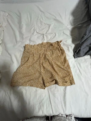 Beige mönstrade shorts med resår - Säljer ett par beige shorts med vitt abstrakt mönster och resår i midjan. De har en lös passform och är perfekta för varma dagar. Materialet känns lätt och luftigt, vilket gör dem sköna att bära hela sommaren.