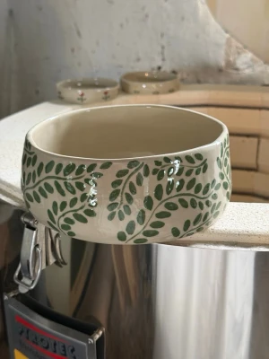 Handmålad keramikskål med bladmotiv - Unik handmålad keramikskål i beige med grönt bladmotiv runt kanten. Skålen har en rund form och blank yta, perfekt som dekoration eller serveringsskål.  Information till dig som köpare: Varje skål är tillverkad för hand, vilket innebär att små variationer och naturliga ojämnheter kan förekomma – det är en del av hantverket och gör varje exemplar helt unikt.  Skålen går att köra i mikro och diska i diskmaskin. Om du vill vara försiktig med den så kör du handdisk💘