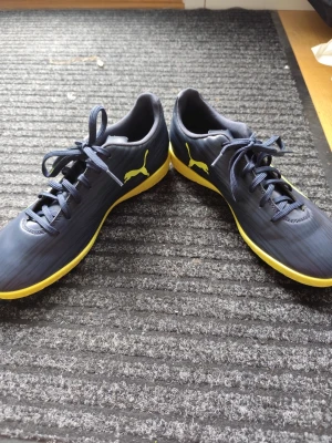 Svarta Puma fotbollsskor med gula detaljer - Snygga fotbollsskor från Puma i svart med gula detaljer och logga på sidan. Skorna har snörning och en låg profil, samt en gul sula som ger extra stilpoäng på planen. Tillverkade i syntetmaterial för bra hållbarhet och passform.