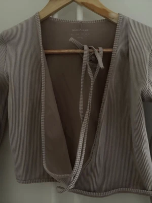 Beige randig omlottopp med knyt - Trendig beige omlottopp från Stradivarius med tunna vita ränder. Toppen har långa ärmar, croppad passform och knyt framtill. Perfekt att styla över ett linne eller topp för en avslappnad look.