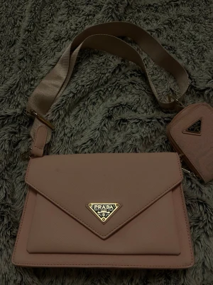 Rosa axelremsväska från Frada - Snygg rosa handväska från Frada med justerbar axelrem och matchande liten väska. Väskan har kuvertformad front med guldfärgad logga och flera fack inuti, inklusive ett med dragkedja. Dock är mini väskan färgad men den går och ta bort, pris kan absolut diskuteras 