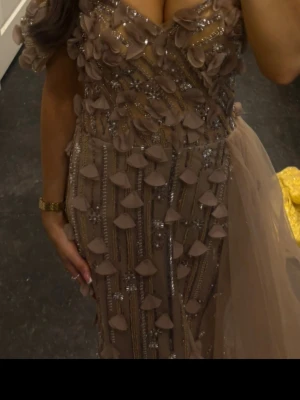 Beige långklänning med paljetter - Elegant beige långklänning med v-ringning, dekorerad med paljetter och 3D-blommor. Klänningen har volangdetaljer och ett skirt släp baktill som ger extra dramatik. Perfekt för dig som vill sticka ut på bal eller fest.