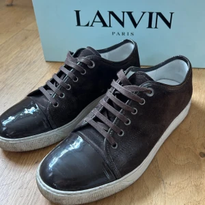 Bruna Lanvins - Snygga bruna sneakers från Lanvin med ovandel i mocka och tåparti i blankt lack. Skorna är i extremt bra skick och knappt använda på 1,5 år. Perfekta för dig som vill ha en clean och exklusiv look. Storlek UK 7 motsvarar 42-43
