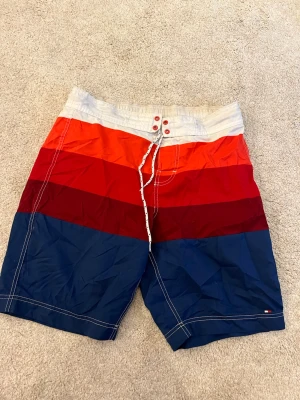 Flerfärgade badshorts från Tommy Hilfiger - Säljer ett par snygga badshorts från Tommy Hilfiger med breda blockränder i vitt, orange, rött, vinrött och marinblått. Klassisk passform med snörning i midjan och bakficka. Perfekta för stranden eller poolen!