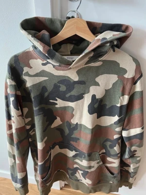 Camo hoodie från WESC - Snygg hoodie från WESC i klassiskt camouflagemönster med gröna, bruna och beige toner. Hoodien har huva och en stor magficka framtill. Tillverkad i mjukt bomullsmaterial som är skönt att bära. Perfekt för en avslappnad streetwear-look. Använd 
