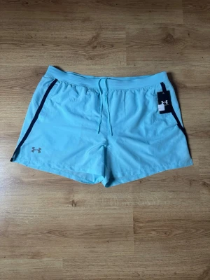 Under Armour - Snygga ljusblå träningsshorts från Under Armour med svarta detaljer längs sidorna och snörning i midjan. Perfekta för gymmet eller löprundan. Tillverkade i lätt och snabbtorkande syntetmaterial perfekt för simmaren. Köpt nya för 449kr, säljer pga för liten storlek. Helt nya och oanvända. Storlek XL, passar M/L.