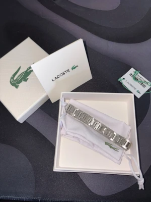 Silverfärgat armband från Lacoste - Stilrent armband i silverfärgat metall från Lacoste med klassisk krokodil-logga. Länkarna har en modern, rektangulär design och armbandet levereras med originalask, påse och tagg. Perfekt accessoar för en trendig look.