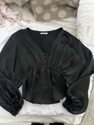 Svart satinblus med v-ringning -  svart blus i glansigt satinmaterial med v-ringning och knappar framtill, resår i midjan och vida, långa ärmar med puff🤗säljer även bikbok vera kostymbyxorna på bilden💕