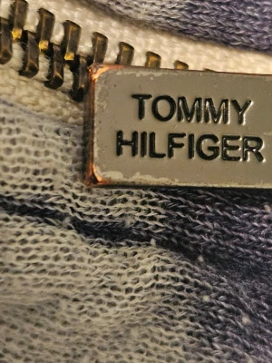 Grå zip-hoodie från Tommy Hilfiger - Säljer en grå hoodie med dragkedja från Tommy Hilfiger. Hoodien har huva med snörning, känguruficka framtill och en liten Hilfiger-logga på fickan. Materialet är mjukt och tvättat bomullstyg för en avslappnad look.