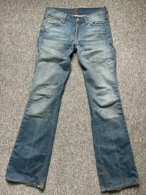 Blå bootcut jeans från 7 for all mankind - Säljer ett par klassiska blå bootcut jeans från 7 for all mankind med slitningar och ljus tvätt. Jeansen har fem fickor, orangea sömmar. Tillverkade i bomull med en avslappnad passform och retrovibbar. Säljer då de blivit för små för mig. 