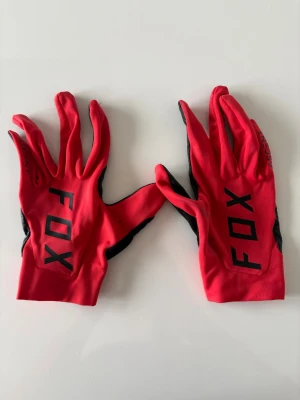 Röda FOX handskar med svart text - Snygga röda handskar från FOX med svart text på ovansidan och svart grepp på handflatan. Perfekta för dig som gillar att sticka ut och vill ha bra grepp. Handskarna har en sportig look och är tunna och smidiga.