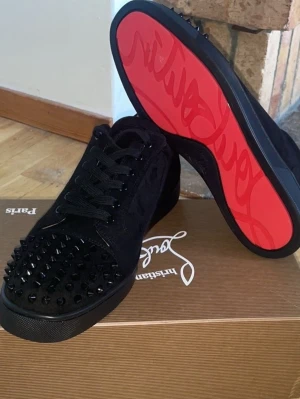 Svarta Louboutin sneakers med nitar - Säljer ett par svarta sneakers från Christian Louboutin med mattsvarta nitar på tån och klassisk röd sula. Skorna är låga och har snörning samt ovandel i mocka för en lyxig känsla. Perfekta för dig som vill sticka ut med en edgy stil.