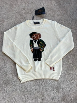 Vit stickad tröja Polo Bear RL - Vit stickad tröja från Polo Ralph Lauren med den ikoniska Polo Bear på bröstet. Tröjan har rund hals, långa ärmar och ribbade muddar. RL-broderi nertill. Perfekt för dig som gillar klassisk och lekfull stil.