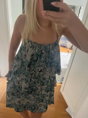 Mönstrad sommarklänning med volanger - Säljer en luftig kort klänning med smala axelband och volangdetaljer. Klänningen har ett abstrakt mönster i blått, grönt och beige, perfekt för varma dagar. Passformen är loose och materialet känns lätt och bekvämt.