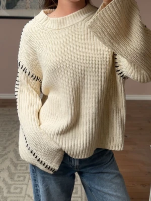 Krämvit oversized stickad tröja - Säljer en krämvit oversized stickad tröja med breda ärmar och svarta dekorativa sömmar längs axlar och ärmar. Tröjan har en ribbad struktur och rund halsringning. Perfekt för dig som gillar en avslappnad och trendig stil.