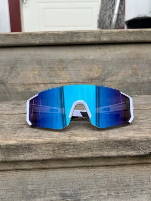 Blå sportiga solglasögon vit ram - Säljer ett par riktigt snygga sportiga solglasögon med spegelblått, stort linsskydd och vit ram. De har en futuristisk, kantig design som passar perfekt för dig som vill sticka ut. Perfekta för utomhusaktiviteter och ger grym stil.
