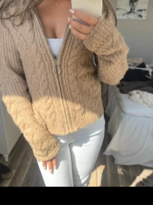 Beige kabelstickad kofta med dragkedja - Mysig beige kabelstickad kofta med dragkedja och ringformad dragkedjetag. Koftan har långa ärmar och en avslappnad passform, perfekt för lager på lager. Stickad struktur ger en trendig look och den neutrala färgen passar till det mesta.
