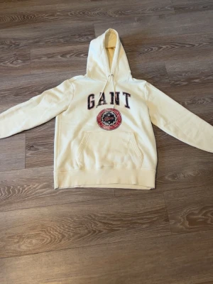 Vit hoodie från GANT med tryck - Snygg vit hoodie från GANT med stor logga och emblem i rött, blått och vitt på bröstet. Klassisk känguruficka framtill och justerbar huva med snören. Tillverkad i mjuk bomullsblandning, perfekt för en avslappnad och sportig stil.
